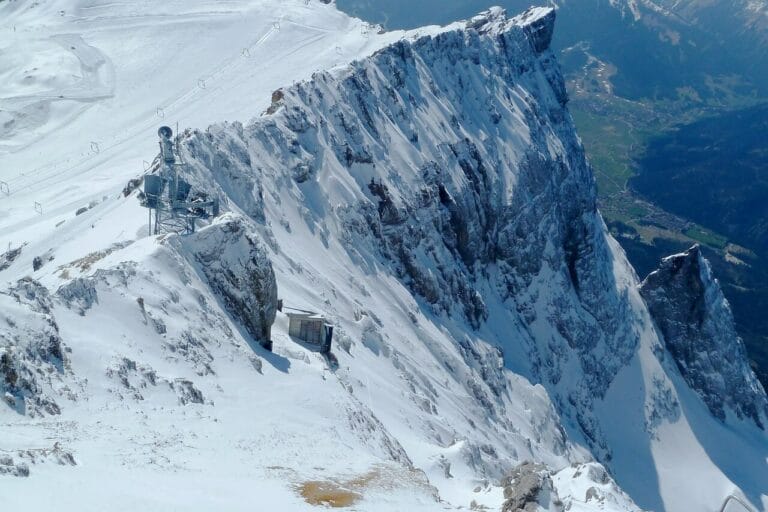 Deutschland höchster Berg Schneebedeckter Berggipfel mit steilen Klippen und entfernten Tälern. Am Hang ist ein kleines Bauwerk zu erkennen, das von eisigem Gelände umgeben ist. Der Himmel ist klar und gibt den Blick frei auf die Berge und Wälder darunter, die in ihrer Erhabenheit an Deutschlands höchsten Berg erinnern.