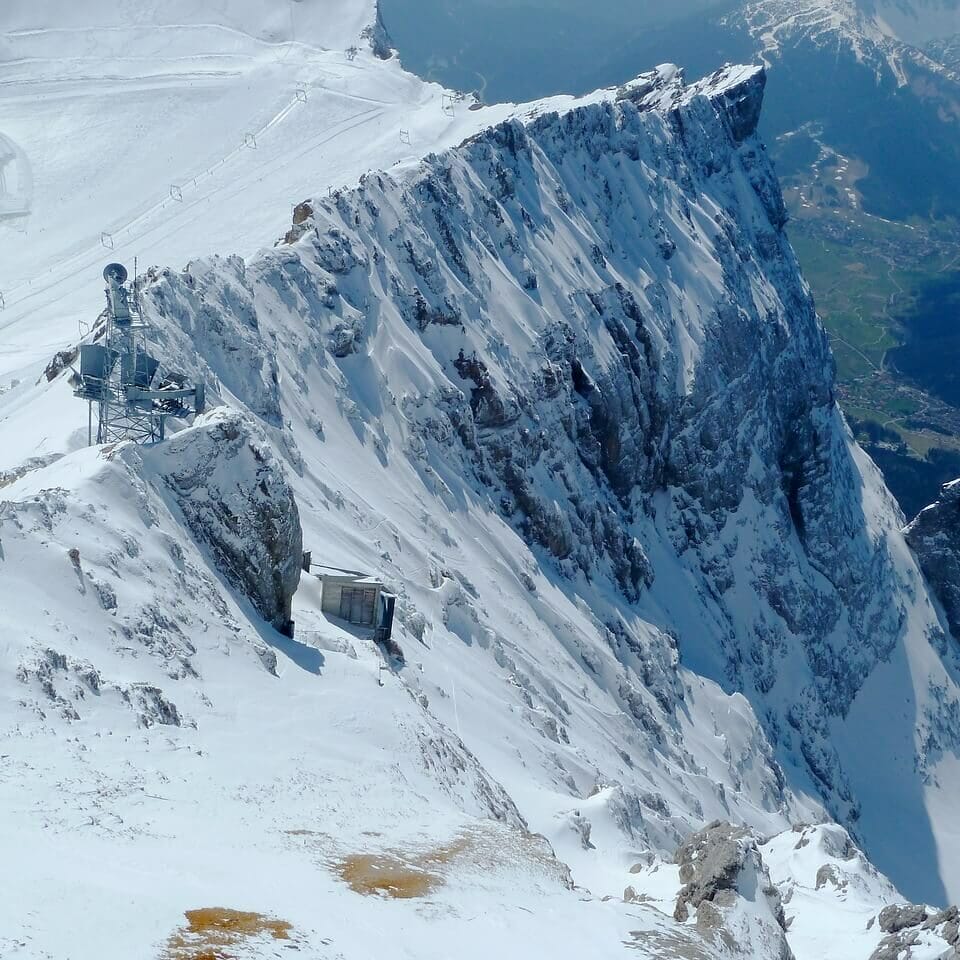 Schneebedeckter Berggipfel mit steilen Klippen und entfernten Tälern. Am Hang ist ein kleines Bauwerk zu erkennen, das von eisigem Gelände umgeben ist. Der Himmel ist klar und gibt den Blick frei auf die Berge und Wälder darunter, die in ihrer Erhabenheit an Deutschlands höchsten Berg erinnern.