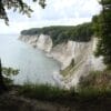 Ein malerischer Blick auf den Königsstuhl auf Rügen offenbart dramatische weiße Kreidefelsen entlang der Küste, gesäumt von üppigen grünen Wäldern. Diese majestätischen Klippen fallen unter einem teilweise bewölkten Himmel steil ins ruhige Meer ab, während Gras und Bäume den Vordergrund wunderschön einrahmen.