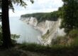 Ein malerischer Blick auf den Königsstuhl auf Rügen offenbart dramatische weiße Kreidefelsen entlang der Küste, gesäumt von üppigen grünen Wäldern. Diese majestätischen Klippen fallen unter einem teilweise bewölkten Himmel steil ins ruhige Meer ab, während Gras und Bäume den Vordergrund wunderschön einrahmen.