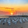 Ein malerischer Strand bei Sonnenuntergang mit Reihen weißer Stühle mit Blick aufs Meer fängt den Reiz der Sehenswürdigkeiten in Mecklenburg-Vorpommern ein. Der Himmel, eine leuchtende Mischung aus Orange und Lila, umrahmt Segelboote in der Nähe eines Wellenbrechers, während üppiges Grün den Sand säumt.