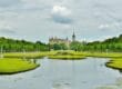 Schloss Schwerin, ein prachtvolles Schloss mit spitzen Türmen, liegt in der Ferne, umgeben von üppigem Grün. Im Vordergrund spiegelt ein ruhiger Teich den bewölkten Himmel wider, gesäumt von gepflegten Rasenflächen und Baumreihen. Statuen schmücken beide Seiten des Wassers.