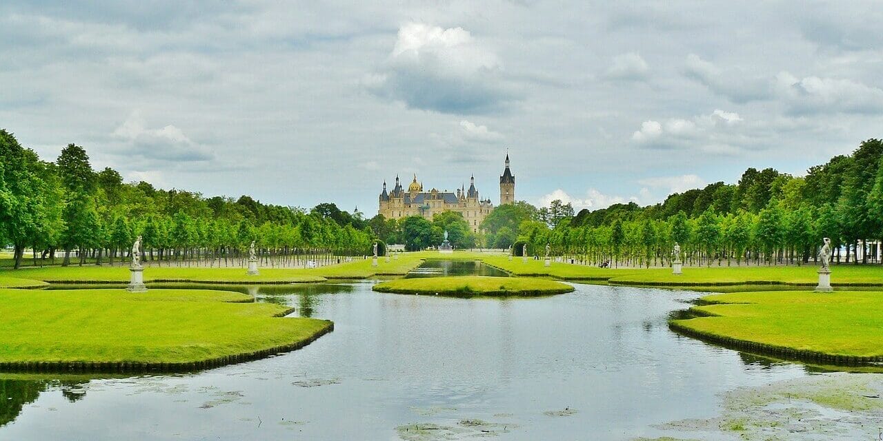 Schloss Schwerin, ein prachtvolles Schloss mit spitzen Türmen, liegt in der Ferne, umgeben von üppigem Grün. Im Vordergrund spiegelt ein ruhiger Teich den bewölkten Himmel wider, gesäumt von gepflegten Rasenflächen und Baumreihen. Statuen schmücken beide Seiten des Wassers.