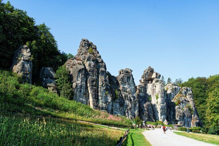 Schönsten Naturwunder in Deutschland Eine Gruppe von Menschen geht auf die Externsteine zu, eines der schönsten Naturwunder Deutschlands, eine Reihe hoch aufragender Sandsteinfelsen, umgeben von üppigem Grün unter einem klaren blauen Himmel.