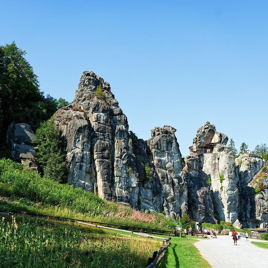 Eine Gruppe von Menschen geht auf die Externsteine zu, eines der schönsten Naturwunder Deutschlands, eine Reihe hoch aufragender Sandsteinfelsen, umgeben von üppigem Grün unter einem klaren blauen Himmel.