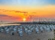 Ein Strand bei Sonnenuntergang in Mecklenburg-Vorpommern besticht durch Reihen weißer Stühle mit Blick auf das Meer. Der Himmel strahlt in Orange-, Rosa- und Lilatönen, während in der Nähe angedockte Segelboote eine ruhige Szenerie schaffen. Perfekte Ausflugsziele für alle, die sanft am Horizont versinkende Sonnenuntergänge zu schätzen wissen.