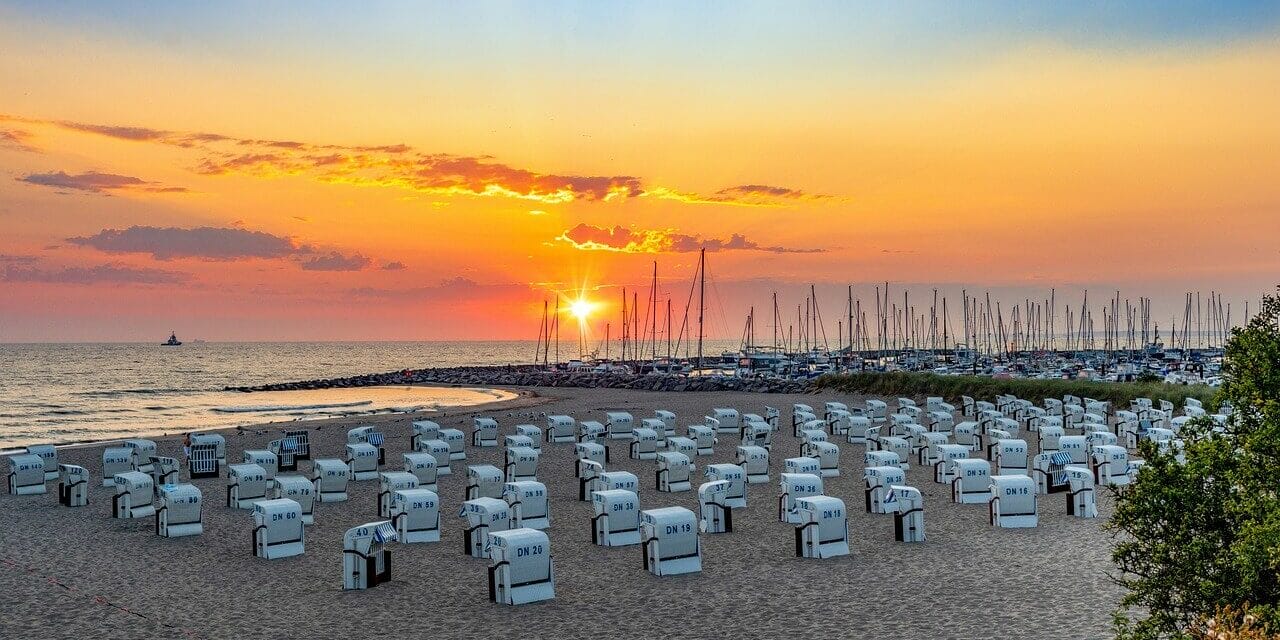 Ausflugsziele in Mecklenburg-Vorpommern Ein Strand bei Sonnenuntergang in Mecklenburg-Vorpommern besticht durch Reihen weißer Stühle mit Blick auf das Meer. Der Himmel strahlt in Orange-, Rosa- und Lilatönen, während in der Nähe angedockte Segelboote eine ruhige Szenerie schaffen. Perfekte Ausflugsziele für alle, die sanft am Horizont versinkende Sonnenuntergänge zu schätzen wissen.