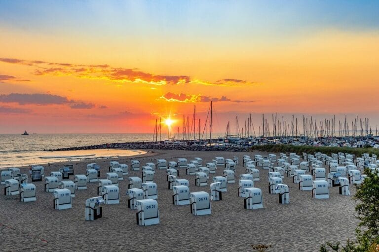 Ausflugsziele in Mecklenburg-Vorpommern Ein Strand bei Sonnenuntergang in Mecklenburg-Vorpommern besticht durch Reihen weißer Stühle mit Blick auf das Meer. Der Himmel strahlt in Orange-, Rosa- und Lilatönen, während in der Nähe angedockte Segelboote eine ruhige Szenerie schaffen. Perfekte Ausflugsziele für alle, die sanft am Horizont versinkende Sonnenuntergänge zu schätzen wissen.