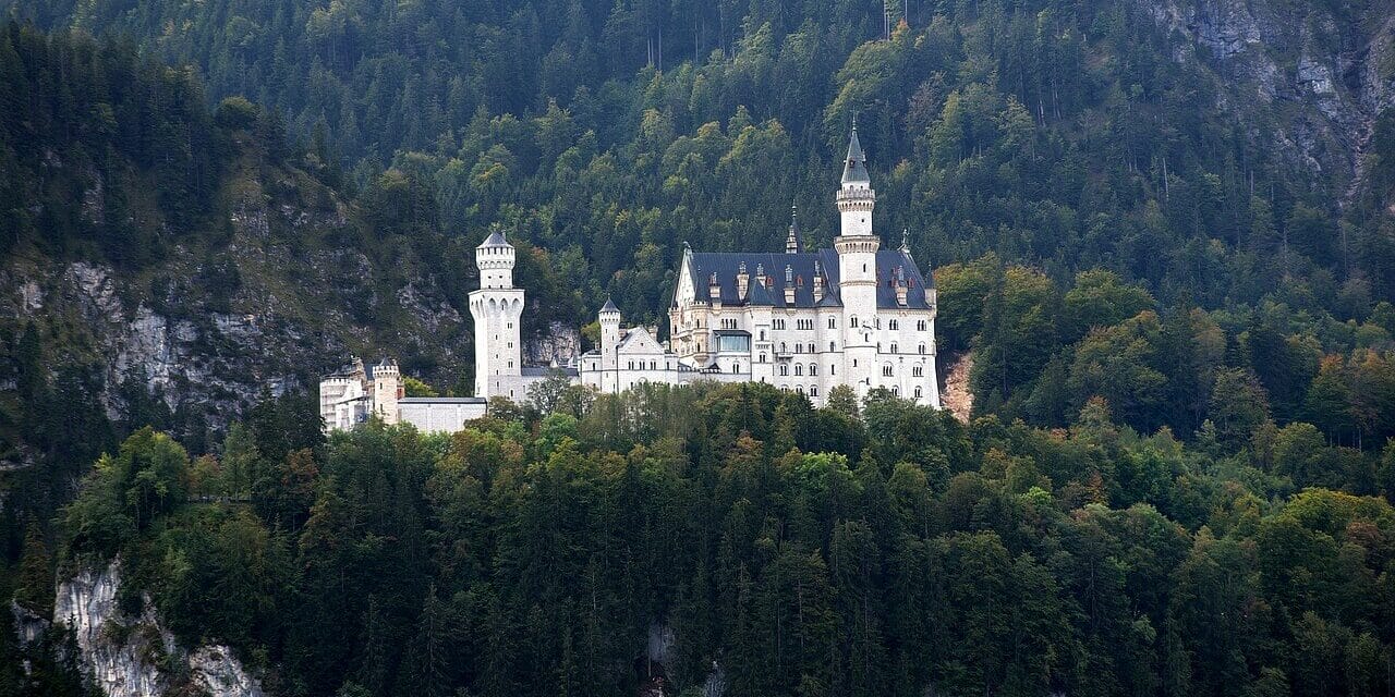 Das malerische Schloss Neuschwanstein liegt auf einem üppig bewaldeten Hügel und besticht durch zahlreiche Türme und Türmchen vor einer Bergkulisse. Umgeben von dichten grünen Bäumen unterstreicht der klare Himmel diese ruhige, märchenhafte Kulisse, die typisch für Burgen und Schlösser in Deutschland ist.