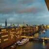 Eine Stadtlandschaft in der Abenddämmerung mit beleuchteten Bürogebäuden entlang eines Kanals zeigt einige der Sehenswürdigkeiten Hamburgs. Boote liegen entlang des Wasserwegs vor Anker und eine Brücke überquert den Kanal. Der bewölkte Himmel fängt einen warmen Schein der Lichter der Stadt ein, die sich im Wasser spiegeln.