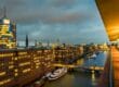 Eine Stadtlandschaft in der Abenddämmerung mit beleuchteten Bürogebäuden entlang eines Kanals zeigt einige der Sehenswürdigkeiten Hamburgs. Boote liegen entlang des Wasserwegs vor Anker und eine Brücke überquert den Kanal. Der bewölkte Himmel fängt einen warmen Schein der Lichter der Stadt ein, die sich im Wasser spiegeln.