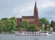Ein weißes Ausflugsboot mit Passagieren fährt auf einem Fluss in der Mecklenburgischen Seenplatte und passiert eine historische rote Backsteinkirche mit einem hohen Kirchturm. Die Kirche ist elegant von üppigen grünen Bäumen unter einem wolkigen Himmel eingerahmt.