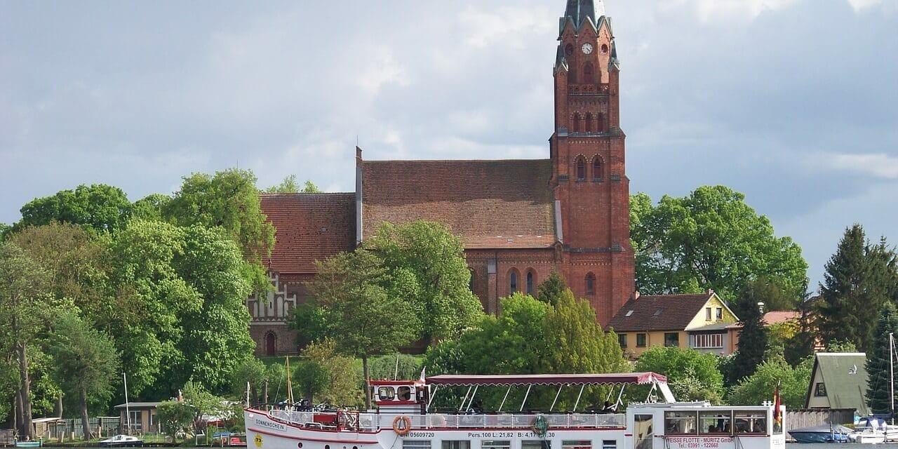 Mecklenburgischen Seenplatte Ein weißes Ausflugsboot mit Passagieren fährt auf einem Fluss in der Mecklenburgischen Seenplatte und passiert eine historische rote Backsteinkirche mit einem hohen Kirchturm. Die Kirche ist elegant von üppigen grünen Bäumen unter einem wolkigen Himmel eingerahmt.
