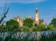 Ein malerischer Blick auf Schloss Schwerin mit seinen verzierten Türmen und Spitzen vor einem klaren blauen Himmel. Im Vordergrund sind üppiges Grün und ein ruhiger See zu sehen, was das märchenhafte Aussehen des Schlosses noch verstärkt und es zu einem der bezauberndsten Schlösser in Mecklenburg-Vorpommern macht.