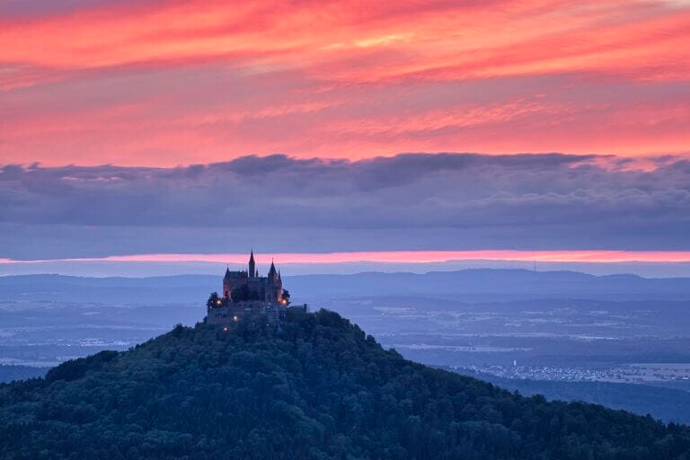 Sehenswürdigkeiten in Baden-Württemberg Ein malerisches Schloss, das an die Burg Hohenzollern erinnert, thront auf einem bewaldeten Hügel und hebt sich vor einem dramatischen Sonnenuntergangshimmel mit leuchtenden Rosa- und Orangetönen ab. Im Hintergrund erstrecken sich sanfte Hügel und ferne Landschaften und schaffen eine ruhige und majestätische Szene, die typisch für Sehenswürdigkeiten in Baden-Württemberg ist.
