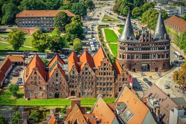 Luftaufnahme von Lübeck, einem Highlight unter den Sehenswürdigkeiten in Schleswig-Holstein, mit mittelalterlicher Architektur mit roten Backsteingebäuden und dem ikonischen Holstentor. Die alten Lagerhäuser und grünen Rasenflächen sind von Straßen und Bäumen unter einem klaren Himmel umgeben.