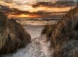 Ein schmaler Sandweg schlängelt sich durch grasbewachsene Dünen zu einem Strand im Sonnenuntergang, der an die ruhigen Strände in Mecklenburg-Vorpommern erinnert. Die Sonne geht über dem Meer unter und wirft warme orangefarbene und goldene Farbtöne in den Himmel und auf die Wolken, während die Wellen sanft das Ufer berühren.